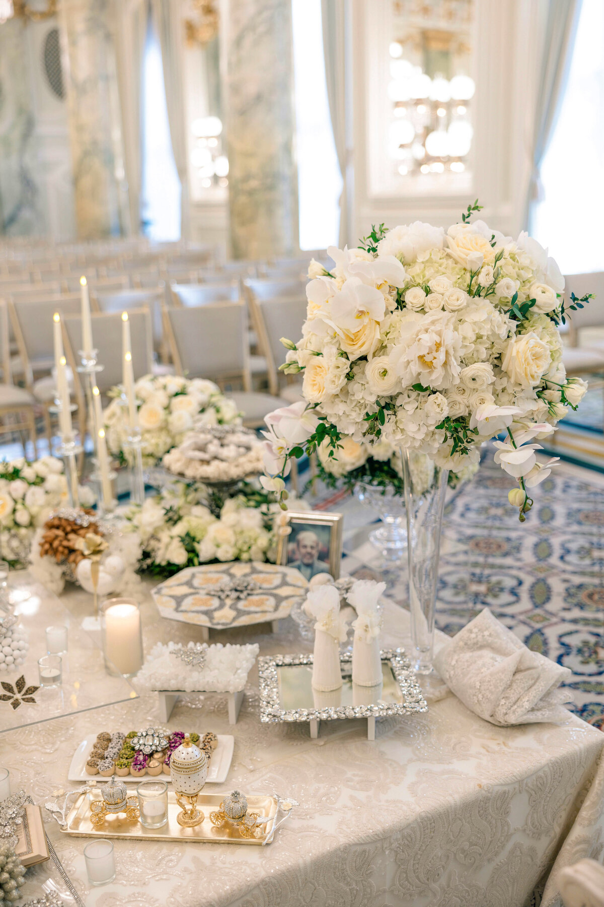 Elegant wedding reception table with ornate white floral arrangements, candles, and decorative platters featuring sweets. The setting includes patterned tablecloths and luxurious decor, creating a sophisticated and romantic atmosphere.