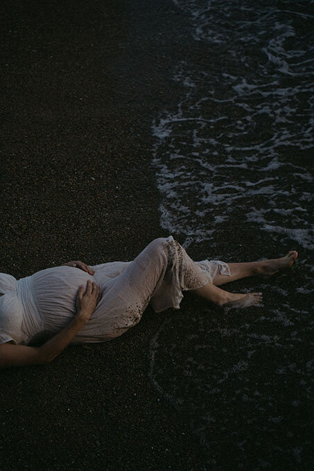 Femme enceinte allongée dans le sable, pieds dans l'eau à la tombée de la nuit. Séance photo grossesse en Bretagne.