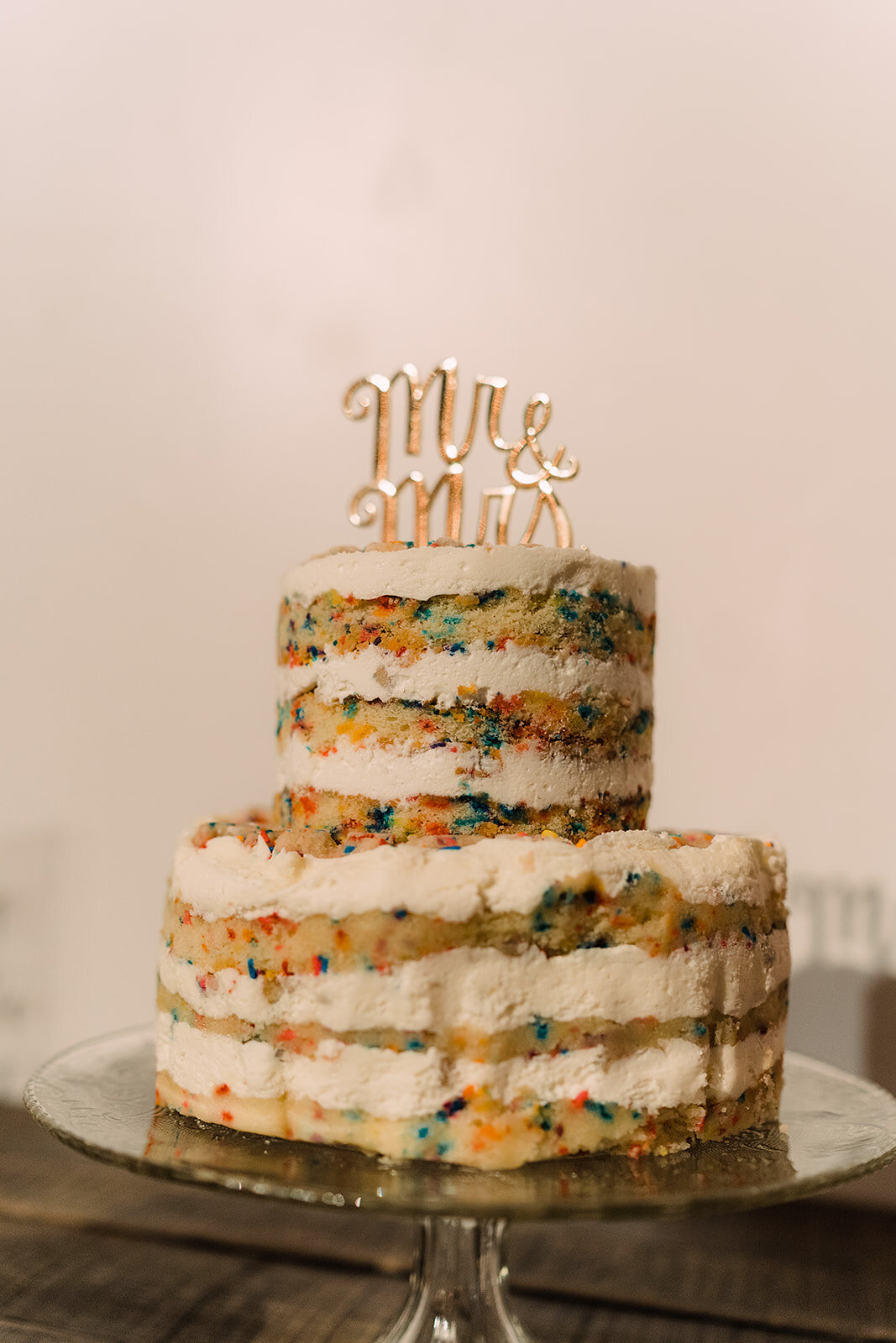 A two-tier naked wedding cake with visible layers of confetti-speckled sponge and buttercream, topped with a golden “Mr & Mrs” sign, adding a whimsical touch.