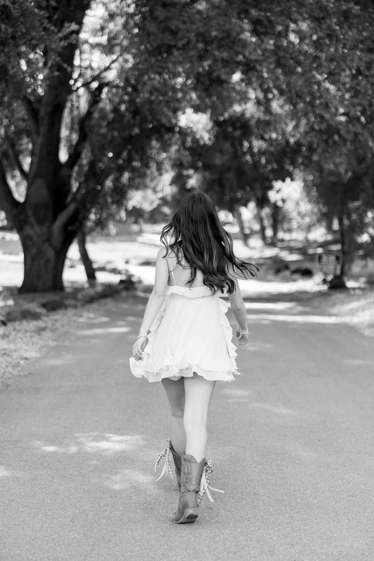 Black and white image of a brunette girl walking away from the camera with a white dress and cowboy boots on