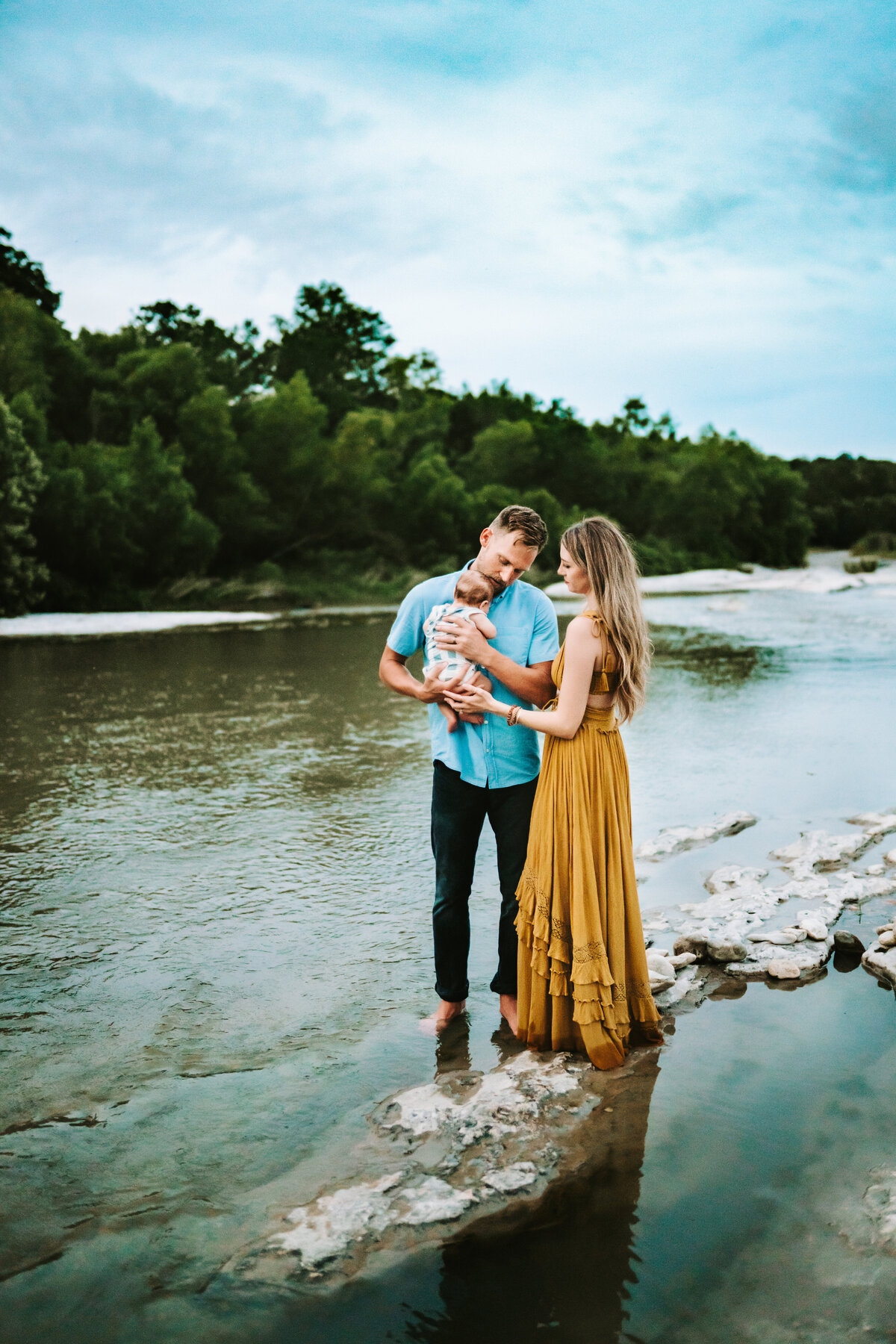 Family Photographer, mom and day stand near a still river admiring their new baby