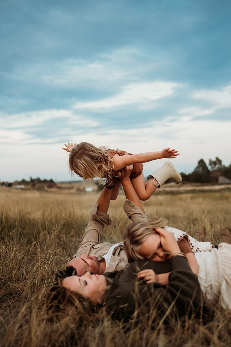 Family  four laying in grassy field on their backs while dad lifts one daughter up into the air.