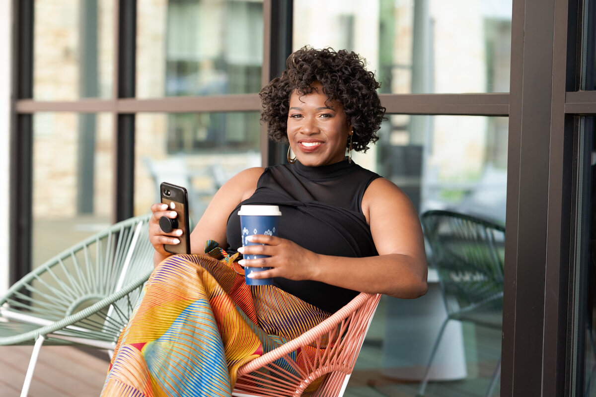 A vibrant and relaxed outdoor portrait of a woman seated on a stylish pink chair, holding a smartphone in one hand and a blue coffee cup in the other. She is dressed in a black sleeveless top paired with a colorful, patterned skirt.