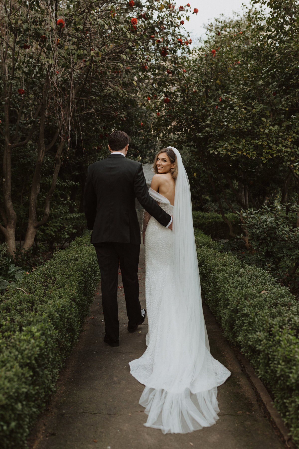 Bride standing with groom in garden pathway while looking back at camera