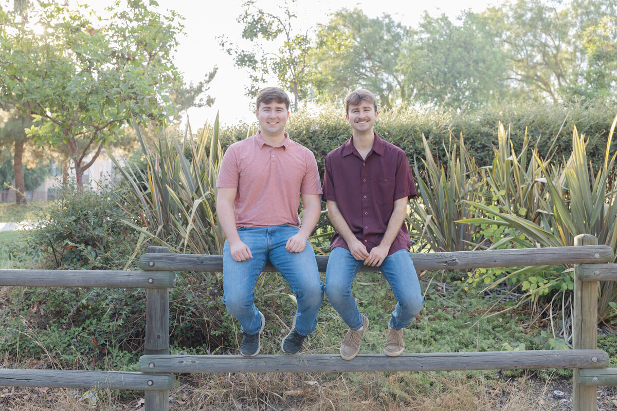 Two brothers wearing blue jeans and shades of red sitting up on a wooden park fence with greenery behind them at Citrus Ranch Park.