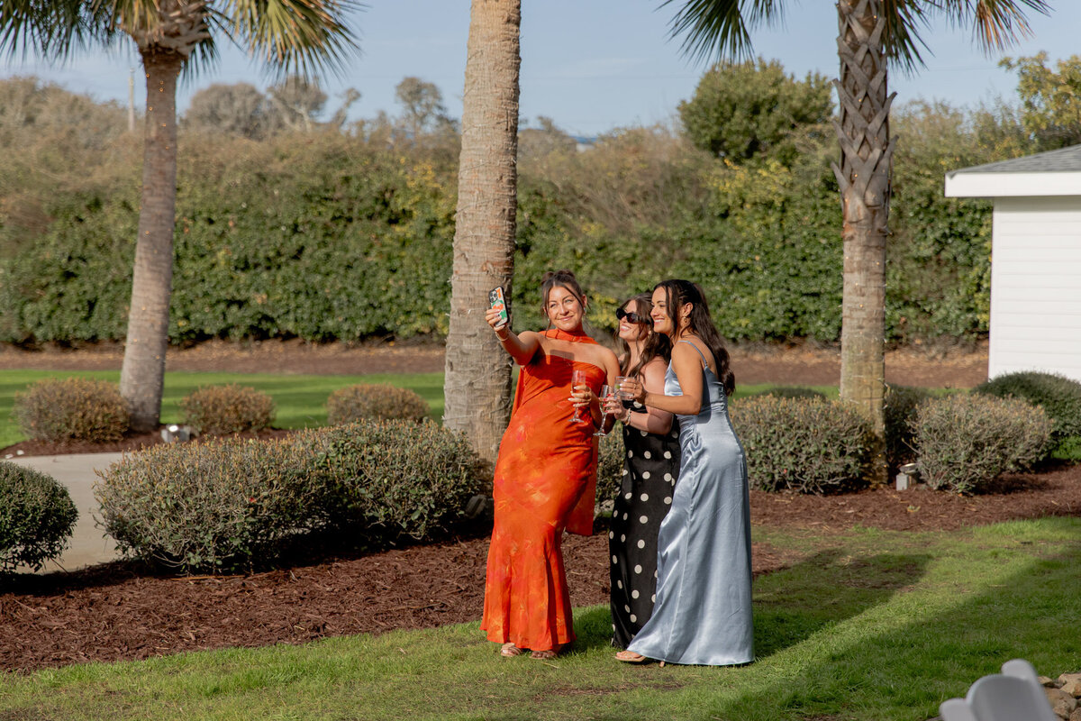 Three girls, all dressed in very different dresses, smile as one of the girls takes a selfie of them. They are outside at an even in Raleigh NC.