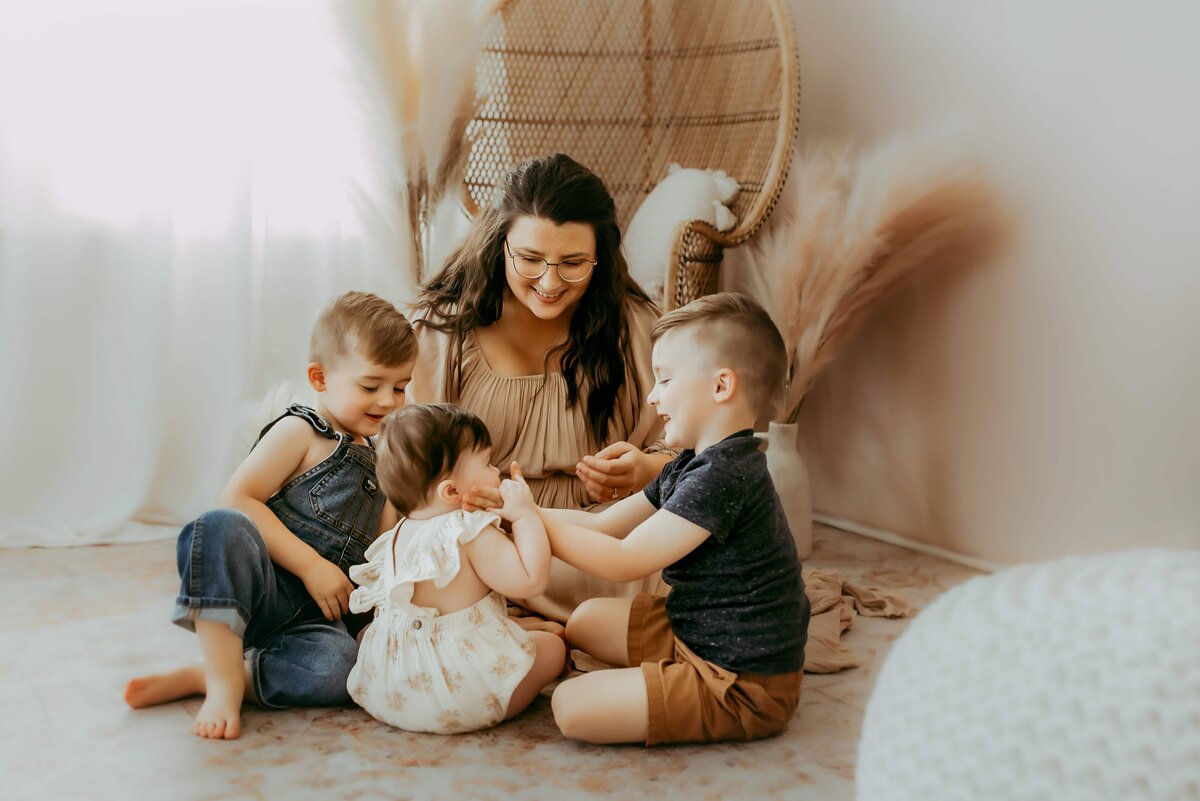 Woman sitting on floor with three children in a softly lit room. Children are playing, and the woman is smiling.