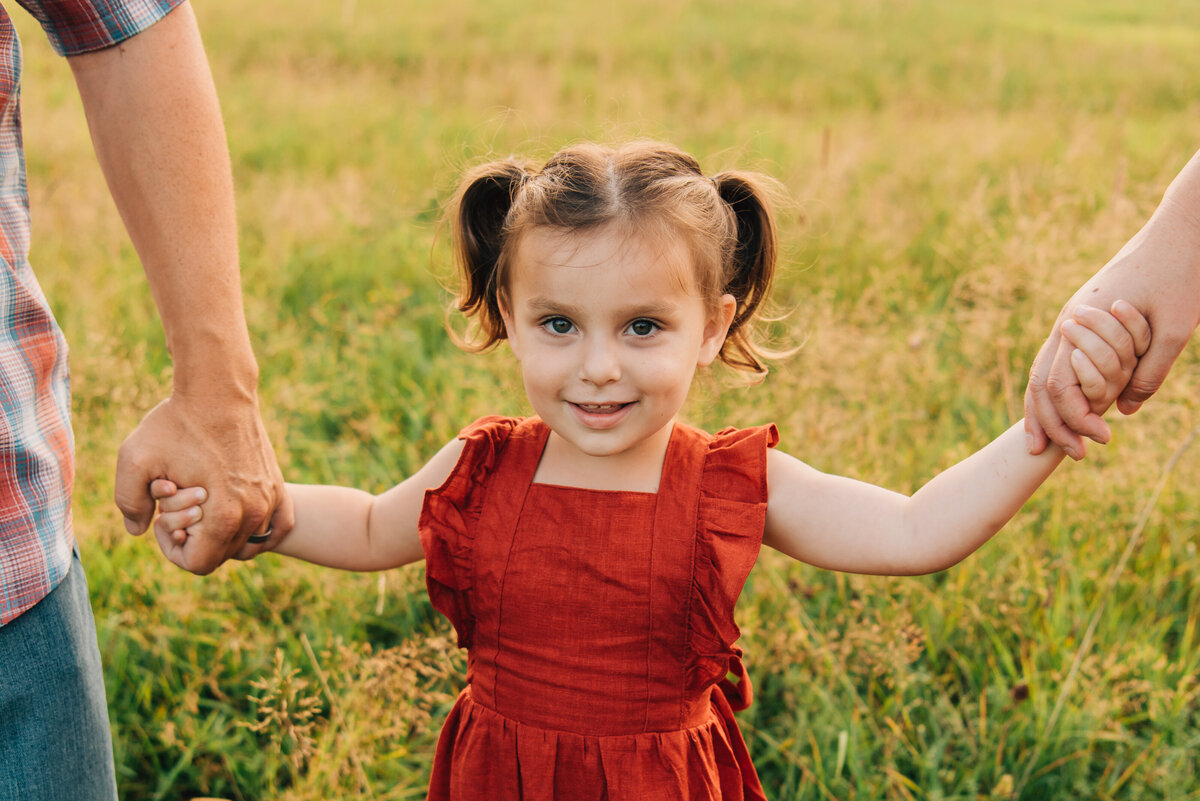 Little girl in red dress smiling at the camera, holding hands with parents