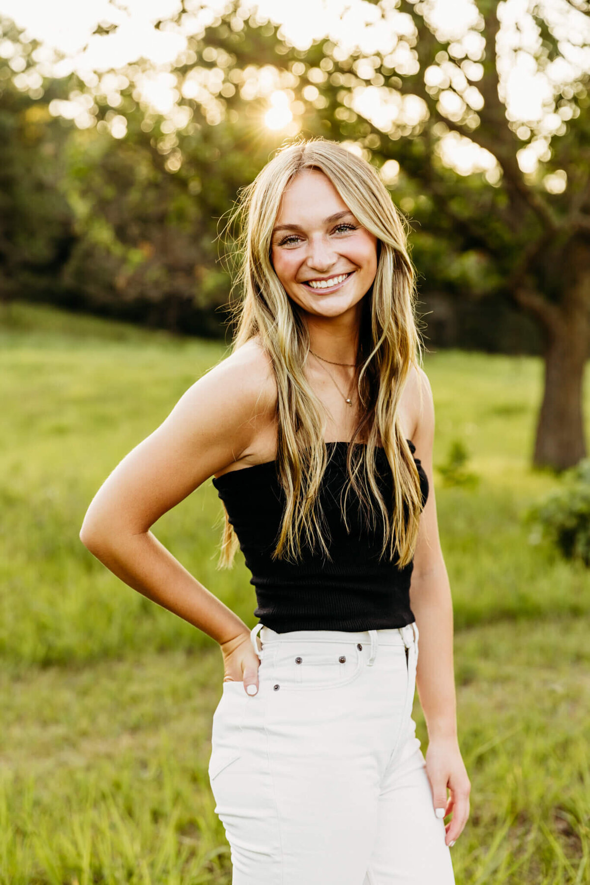 pretty teenage girl in a black top and white jeans standing with one hand in her back pocket as she smiles for her senior photos
