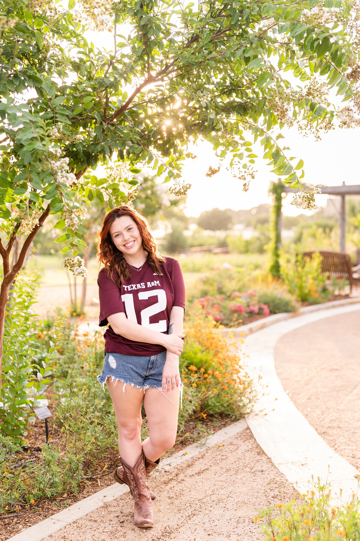 Texas A&M senior girl holding elbow and smiling while wearing maroon jersey and boots in Leach Teaching Gardens
