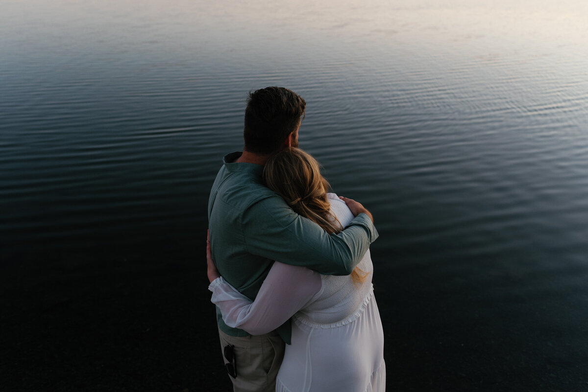 Couple hugging in front of calm water at sunset.