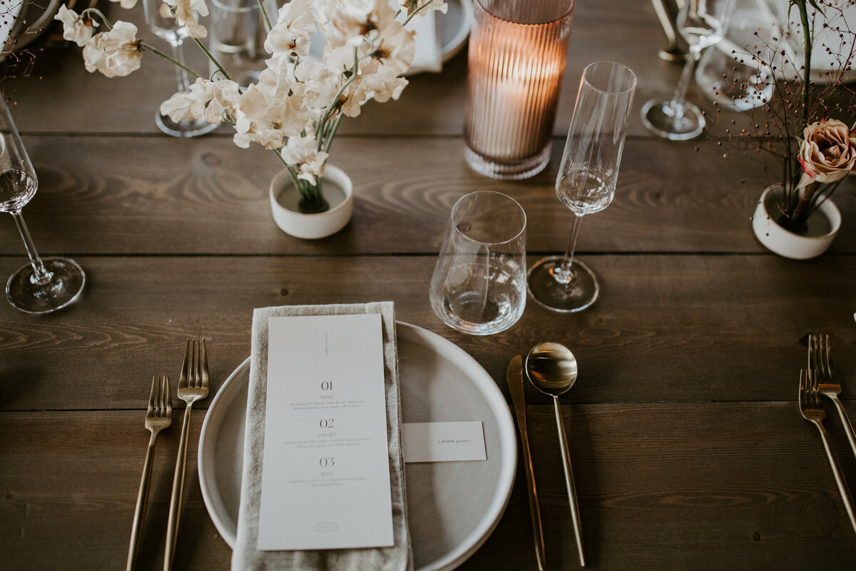 White dinner menu with black font atop a white linen napkin and white plate set on a wooden table with glassware and flowers.