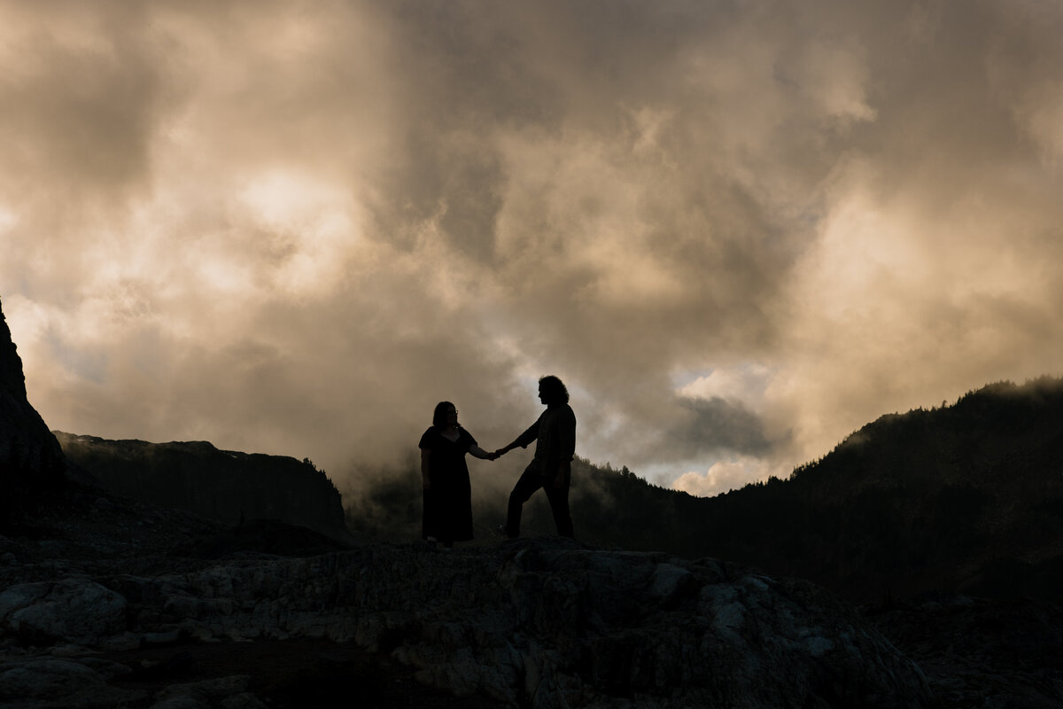 A silloutte of a couple holding hands on a cliffside with dramatic clouds above them