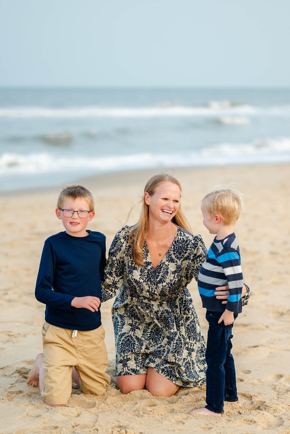 A mom kneels on the beach with her sons. She is laughing at something the younger son said to her.
