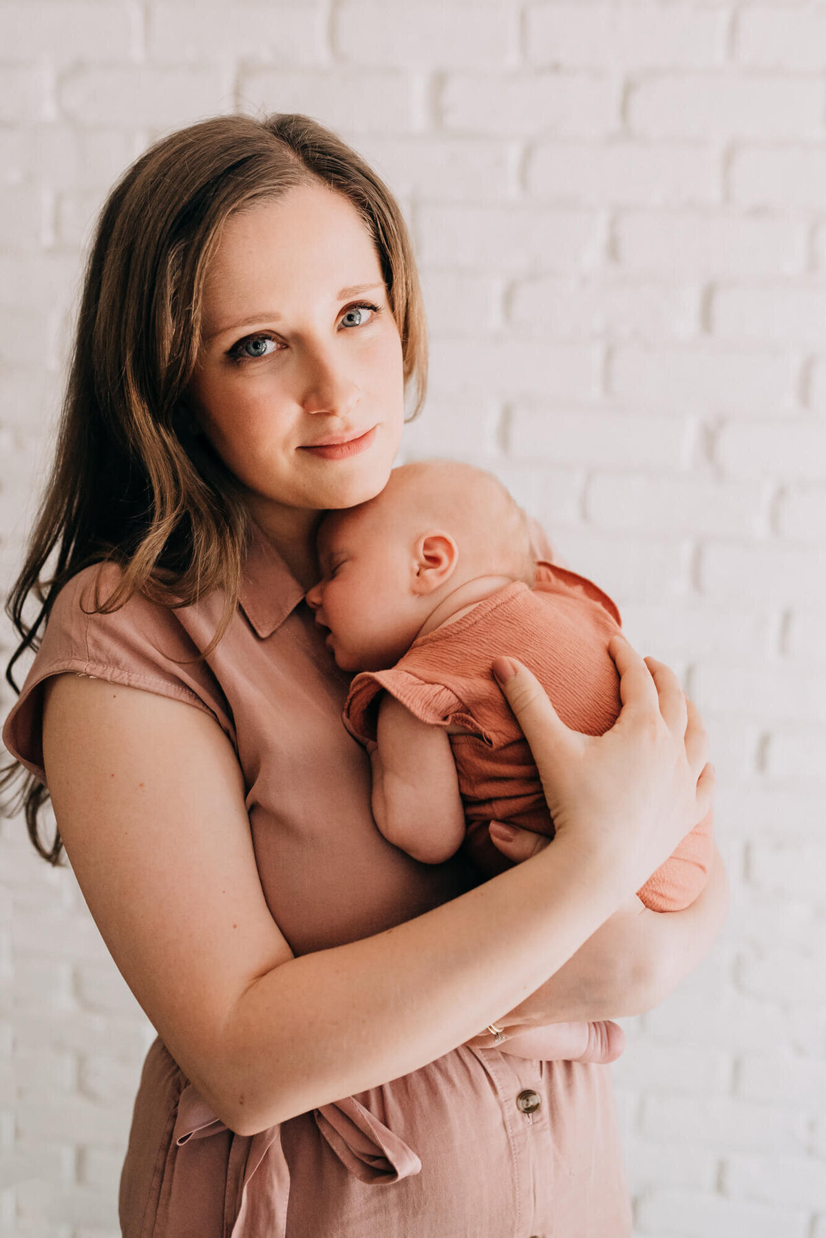 A young mother looks at the camera while holding her newborn daughter in front of a white brick backdrop