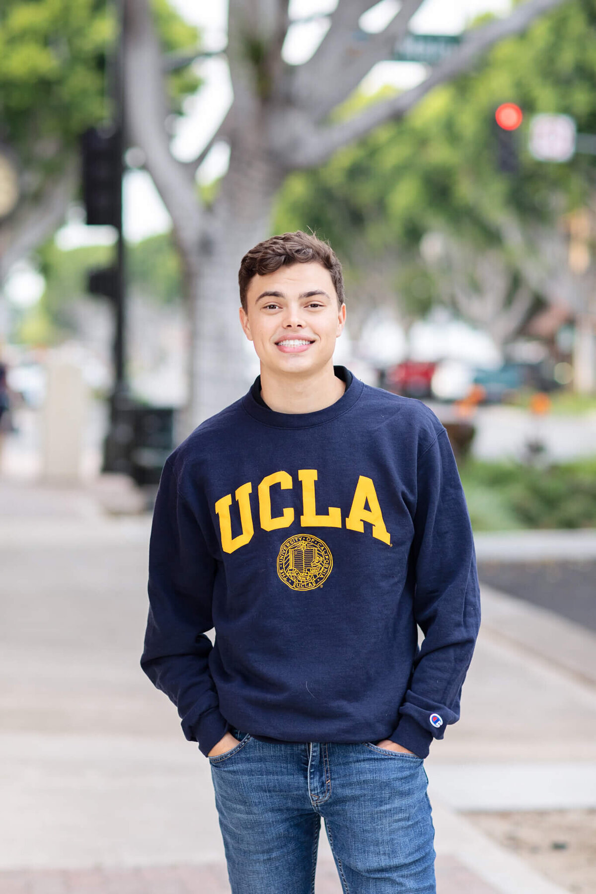 A person wearing a navy UCLA sweatshirt stands outdoors on a sidewalk with blurred trees and street in the background. They have short hair and are smiling, with hands in jean pockets.