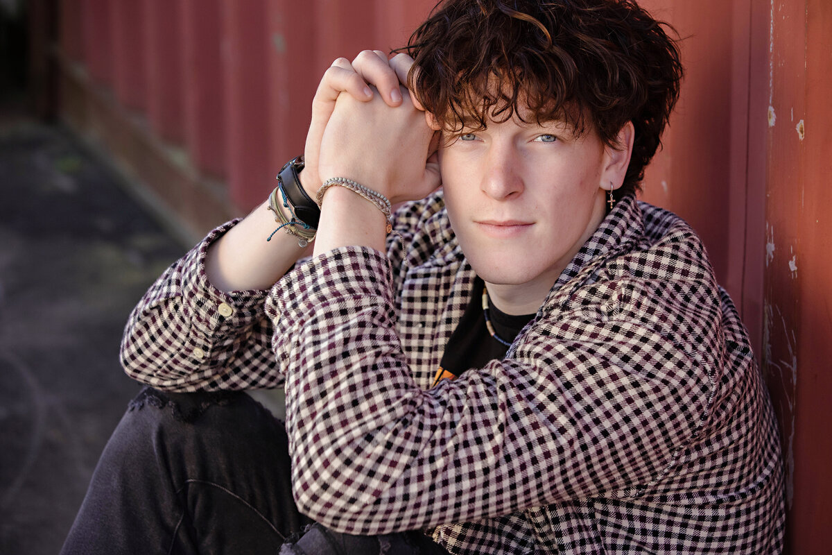 Seated senior portrait in front of a red cargo container at Camp North End, wearing a Beatles tee and checked shirt, captured by Charlotte portrait photographer.