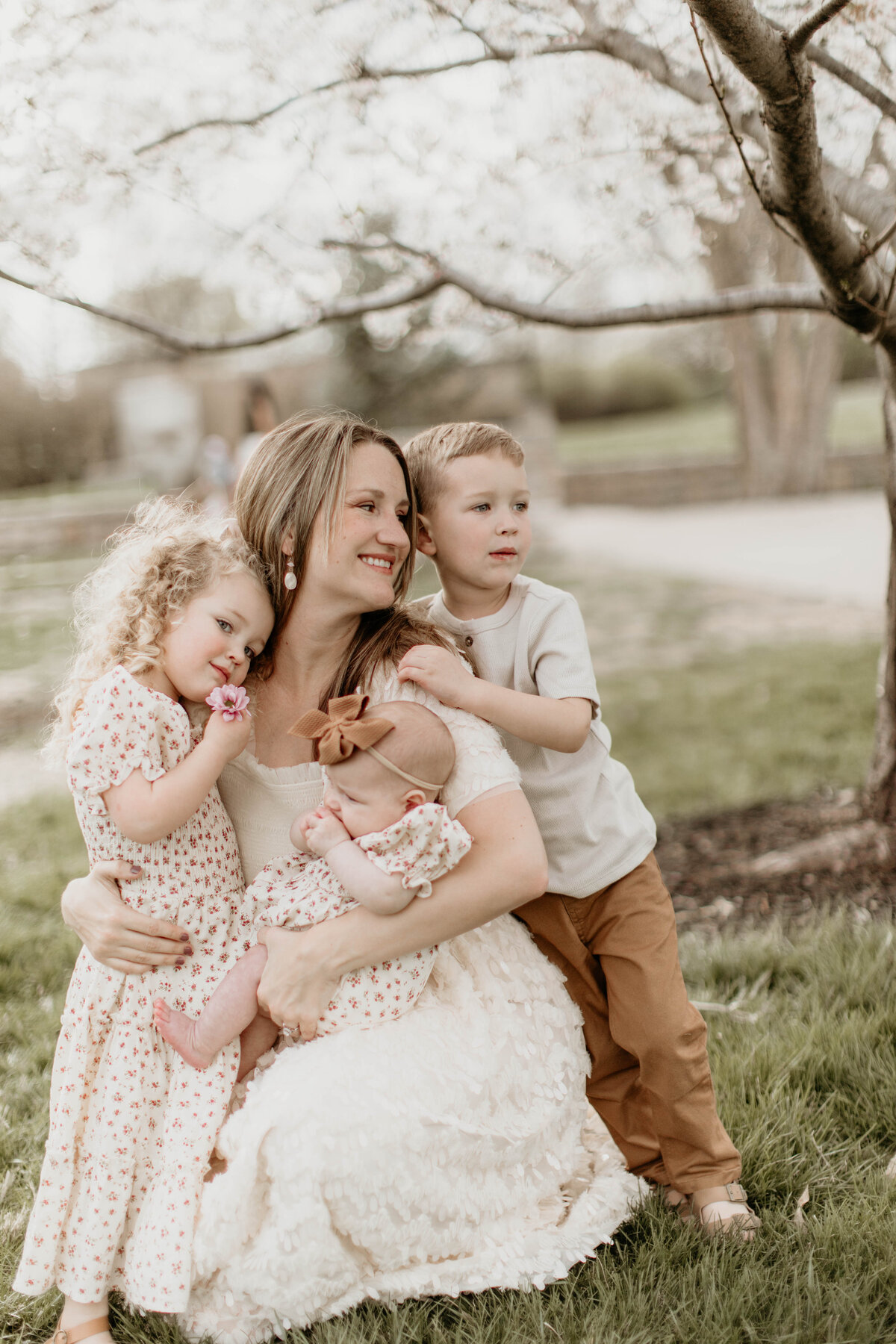 Mom snuggling all three kids in her lap under a tree, soft light surrounding them.
