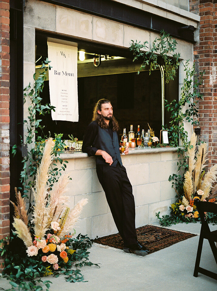 Groom wearing a black suit leans against the bar outside, surrounded by flowers and green garland.