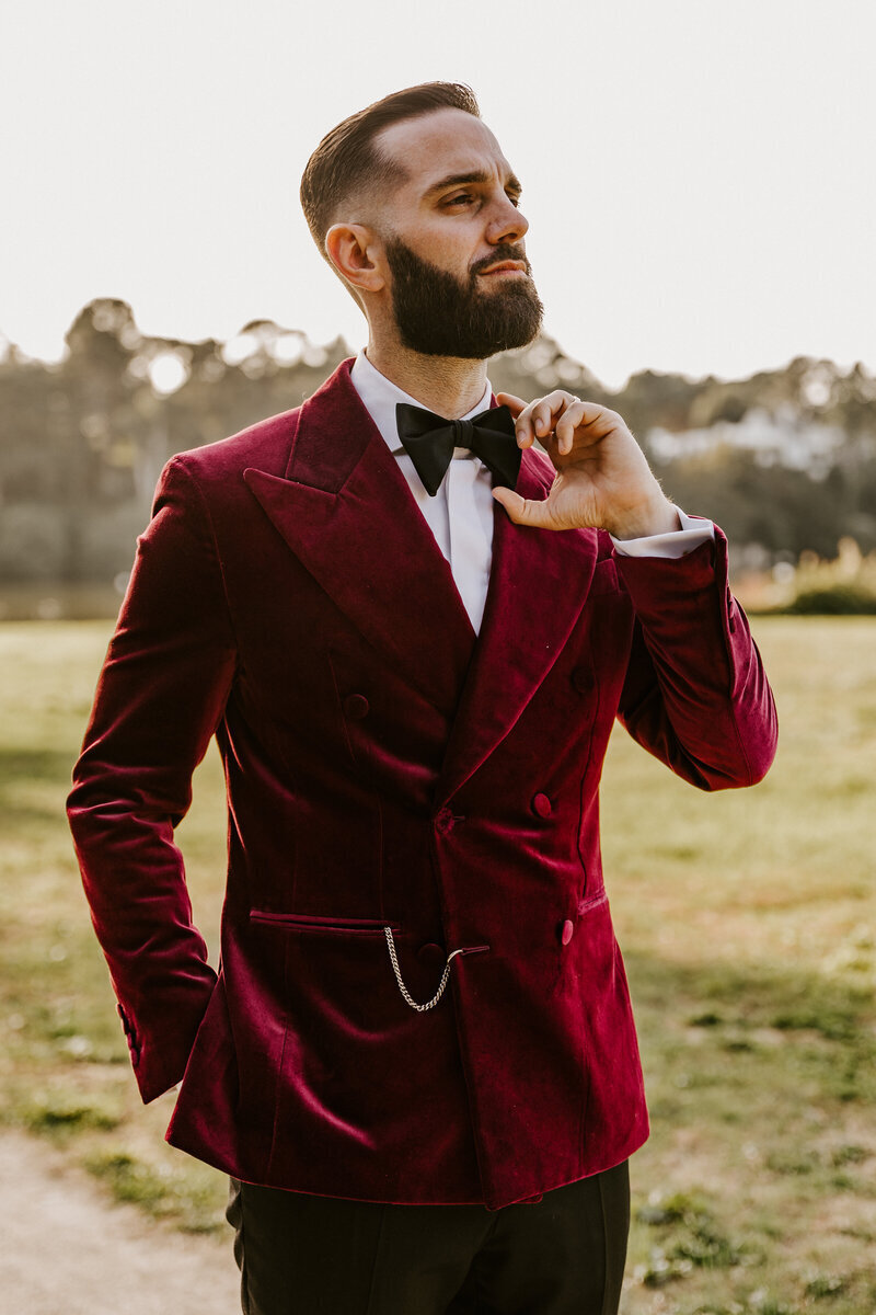 Marié en costume de velours rouge, main droite dans la poche et main gauche ajustant le noeud papillon dans un cadre extérieur, posant pour une séance photo mariage en Vendée.