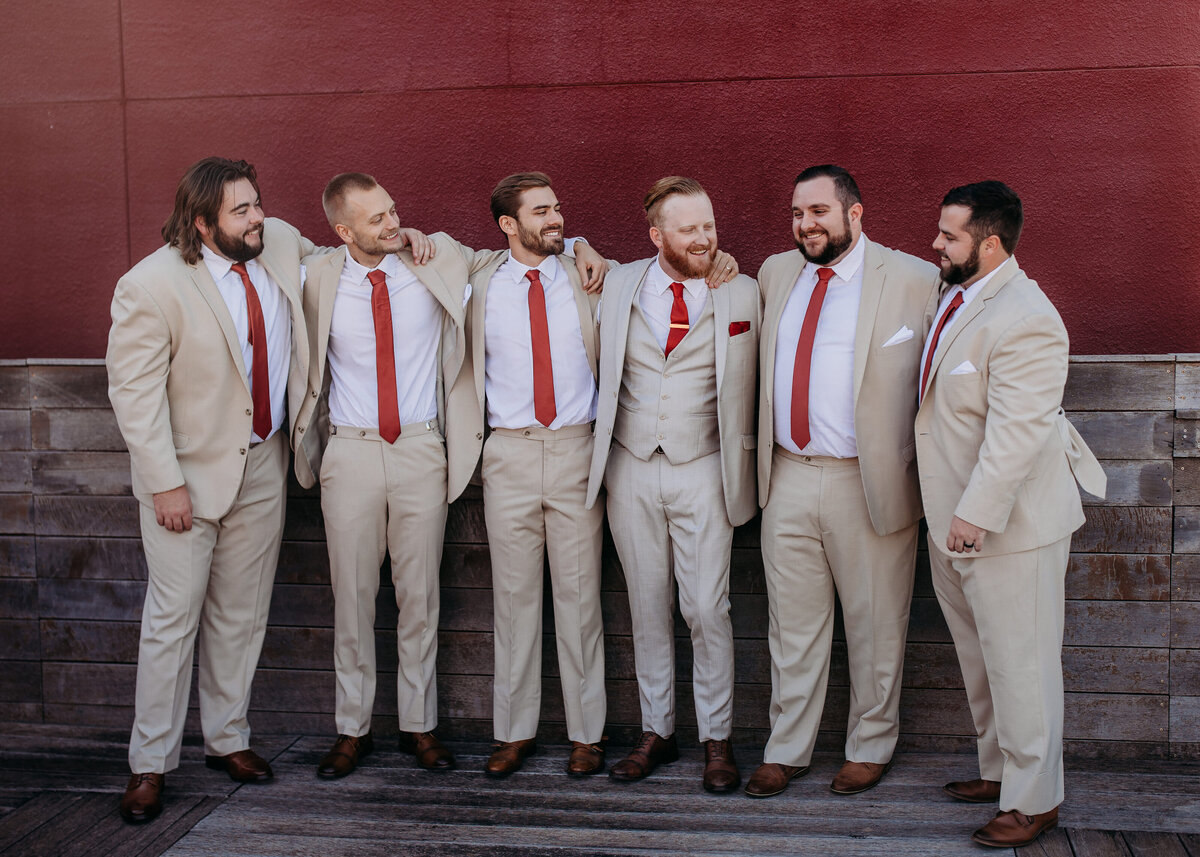 Group of groomsmen lined up against a red wall, all in beige suits with red ties