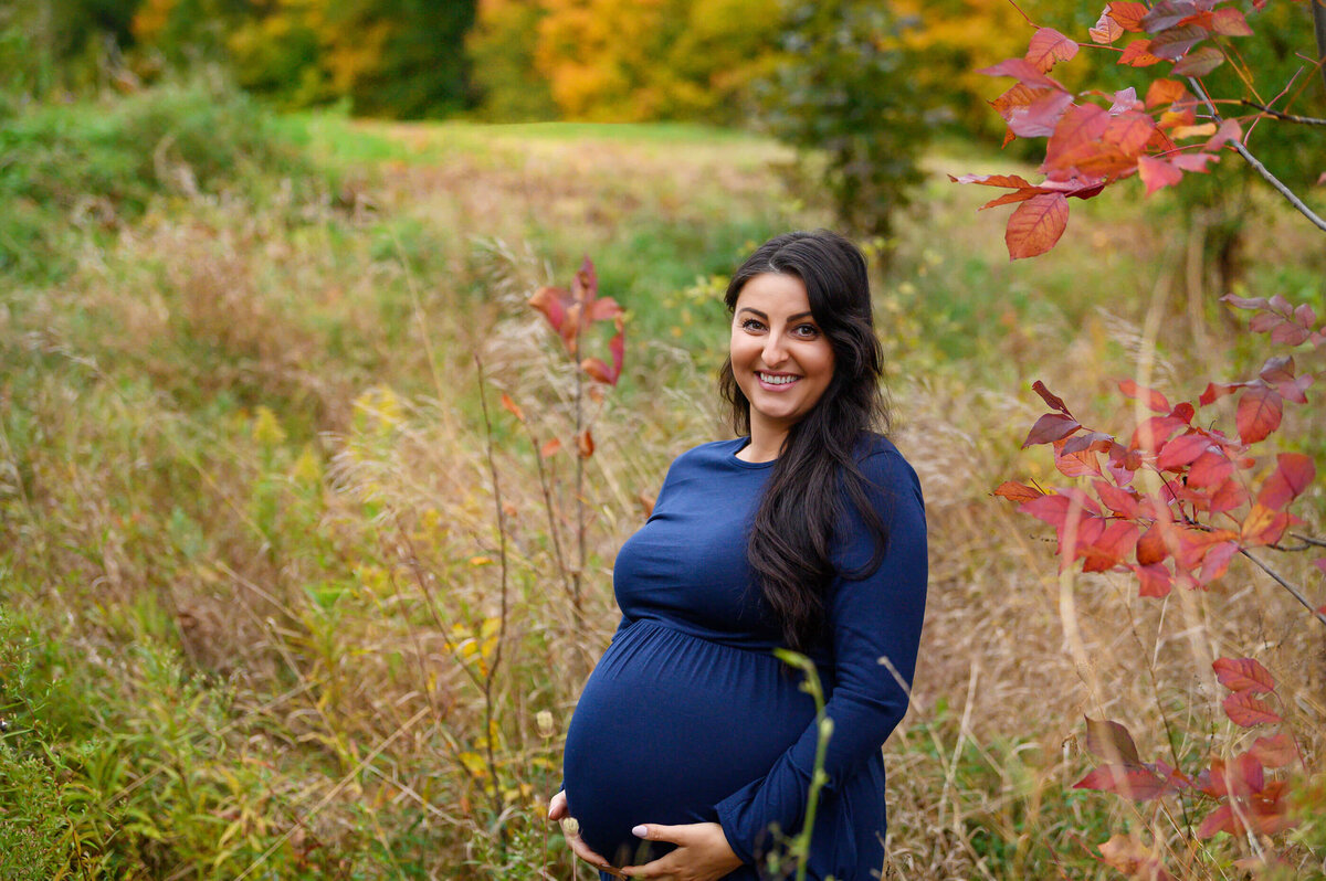 pregnant woman wearing a blue maternity dress in a field with fall foliage for her outdoor maternity photography session in Burlington, Ontario