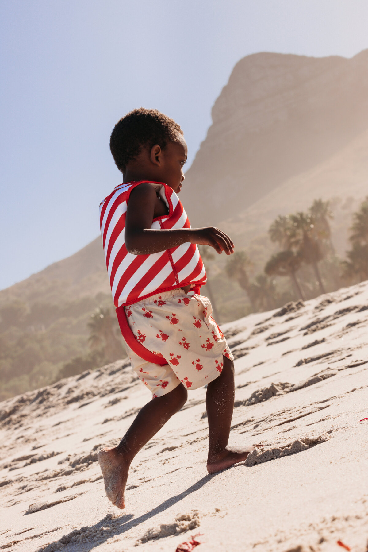 Young boy in a red-and-white striped swim vest and crab-print shorts walking uphill on a sunny beach with palm trees and a mountain in the background in Cape Town.