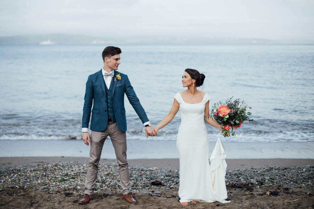Devon Wedding Photographer stood on the beach in a wedding dress holding hands with her husband