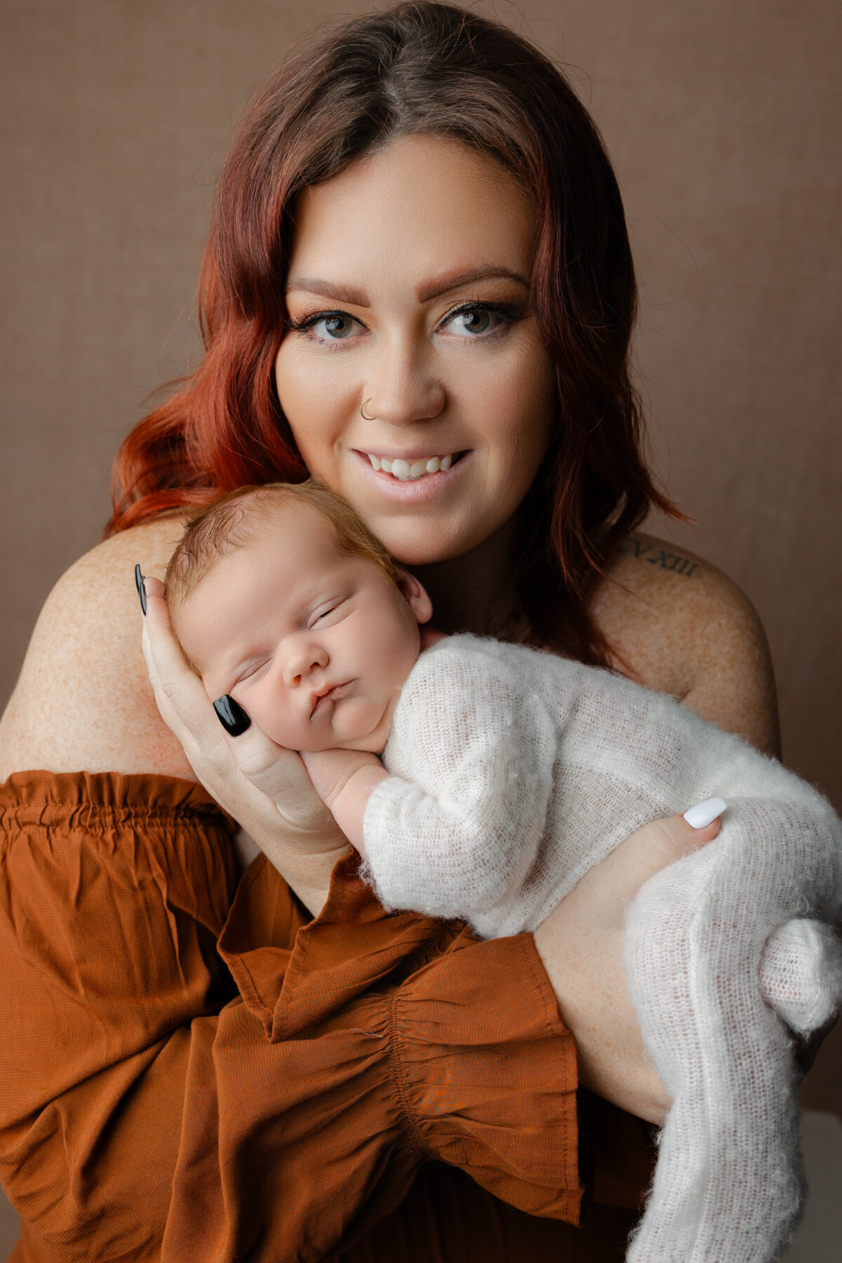 Mother smiling while holding her newborn baby girl, who is swaddled in a white knit outfit and resting peacefully in her hands. Captured by Inland Empire newborn photographer Kaitlyn Dawn Photography.