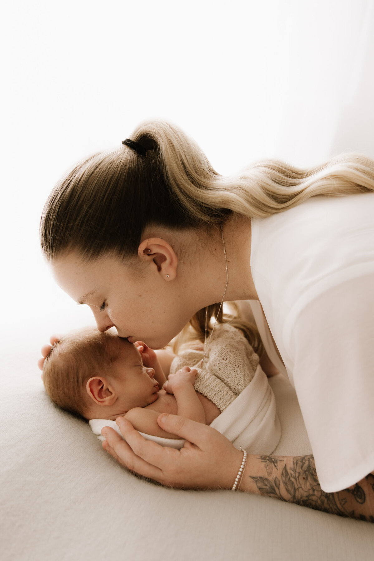 Mother gently kisses her newborn baby’s forehead while holding them closely on a soft cream blanket. The baby is swaddled and dressed in a delicate knit outfit. Captured in a Sydney newborn photography studio with soft diffused lighting, the image conveys tenderness, protection, and deep maternal love.