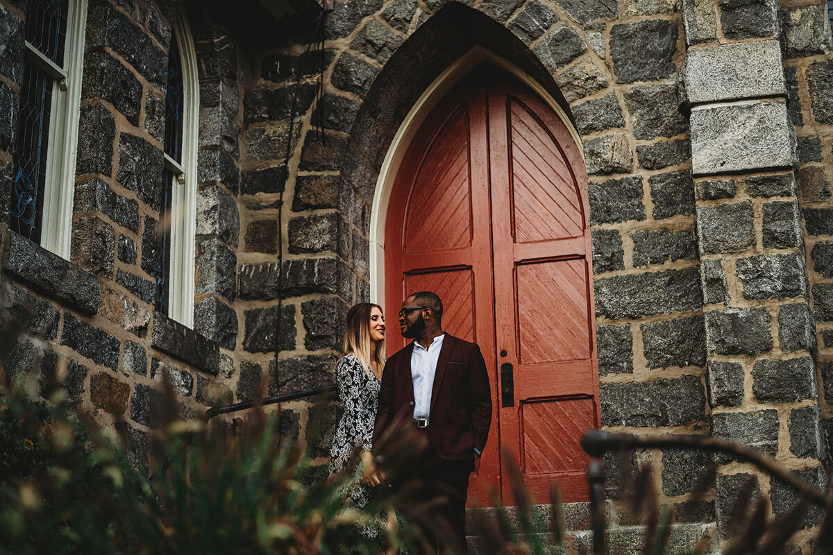 Baltimore wedding photographers captures outdoor engagement session with man and woman holding hands outside of a old stone building with a large red door