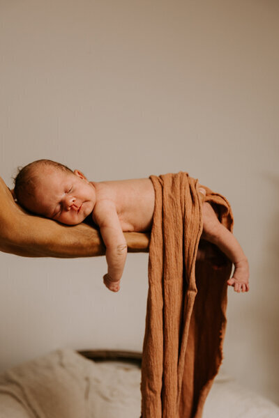 Nouveau né endormi recouvert d'un tissu, reposant sur l'avant-bras de son papa lors d'un shooting photo naissance.