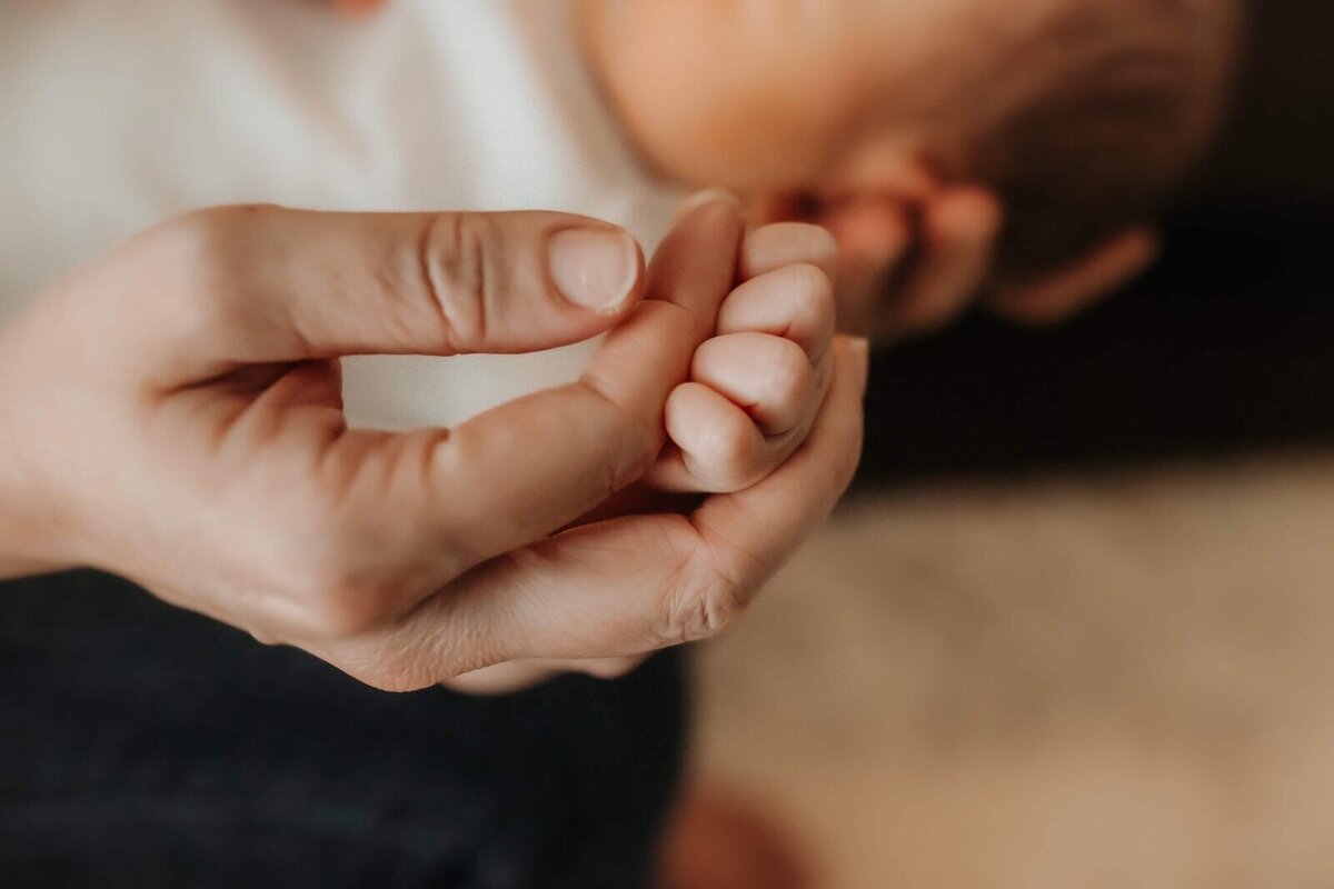An adult hand gently holds a baby's tiny hand, with both hands in soft focus.