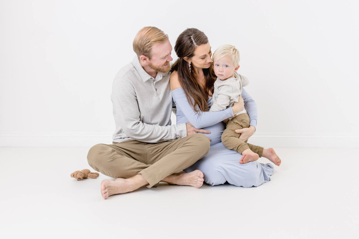 A family sits together on the floor against a white background. A father and a mother, with long hair, hold their young child. A small teddy bear lies nearby. The parents look at the child lovingly.
