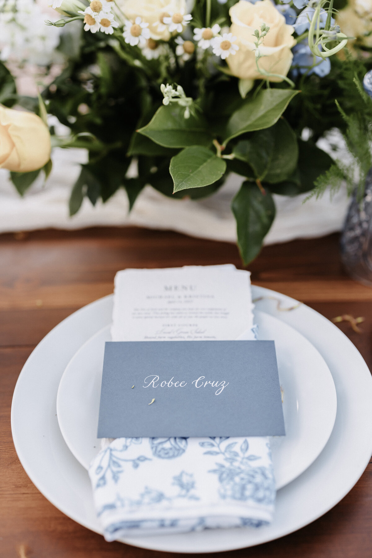Close up of wedding reception napkin and plates wtih centerpiece in the backgrond and name card that reads Robee Grey