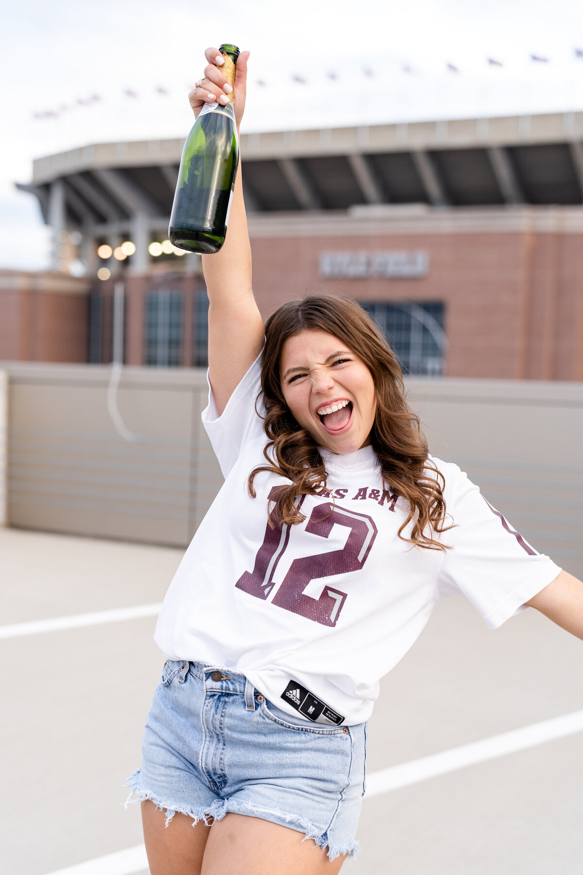 Texas A&M senior girl celebrating with champagne bottle wearing white jersey on rooftop garage in front of Kyle Field