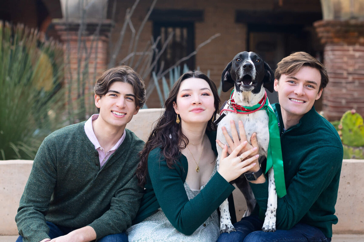 Three siblings with dark hair, dressed in shades of green smiling while holding their black and white spotted dog.