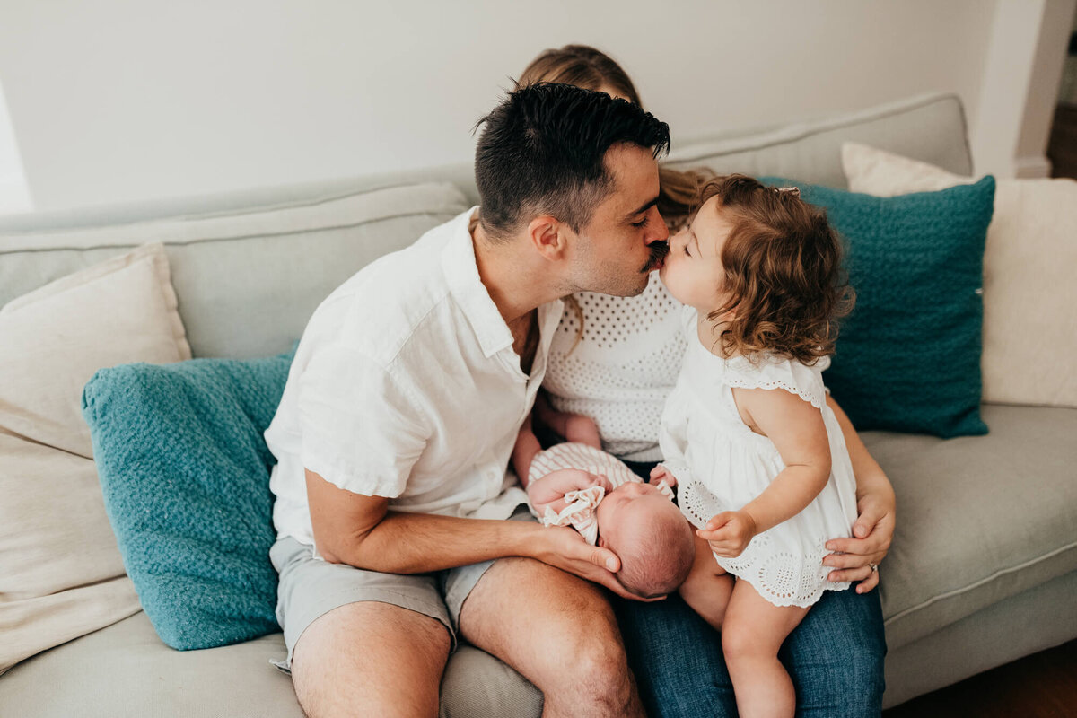 A little girl gives her father a kiss while he holds her baby sister on their couch
