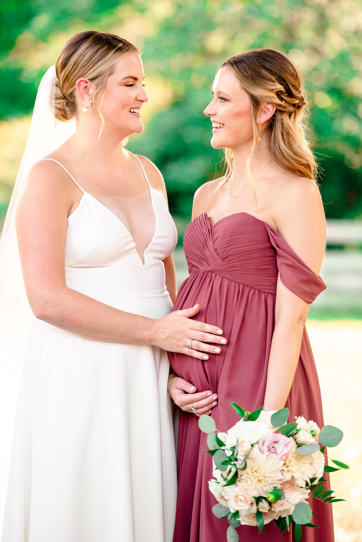 Two women stand smiling at each other outdoors. One is in a white wedding dress with a veil, and the other in a mauve gown holding a bouquet and gently touching her baby bump. The background is a sunlit, blurry garden.
