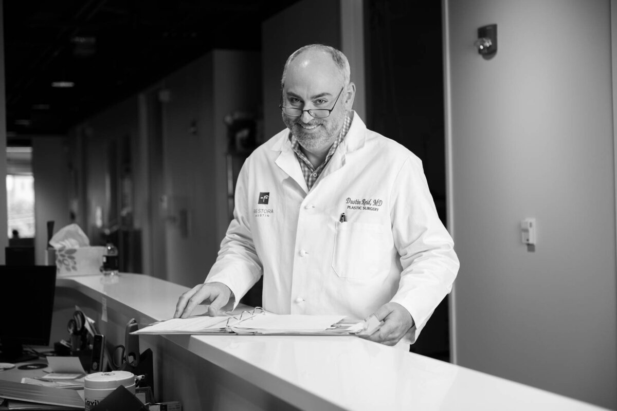 A black and white image of a man wearing glasses and a white coat standing in a clinic