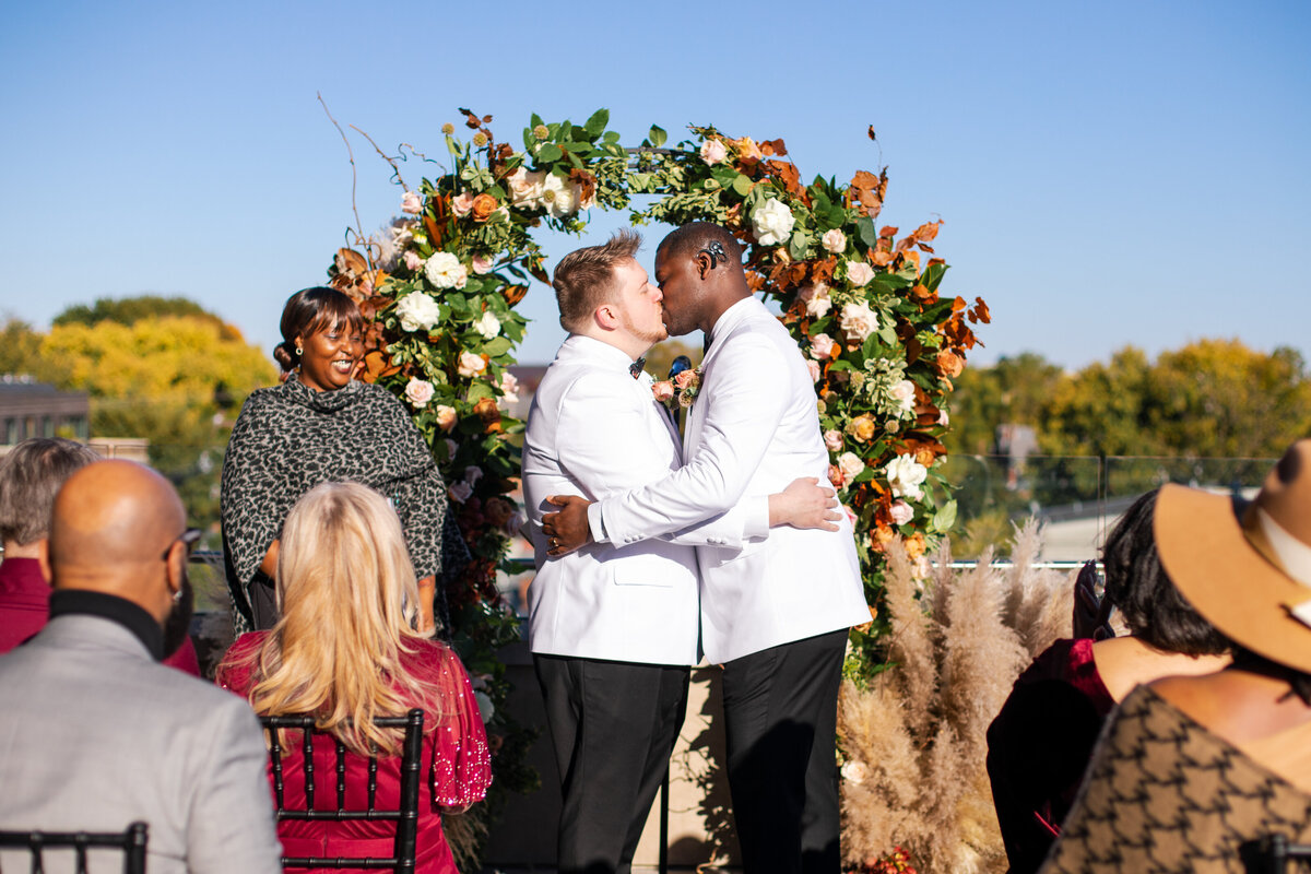 Two grooms share their first kiss in front of a colorful arch at their rooftop wedding at The Terrace in Columbus, Ohio.