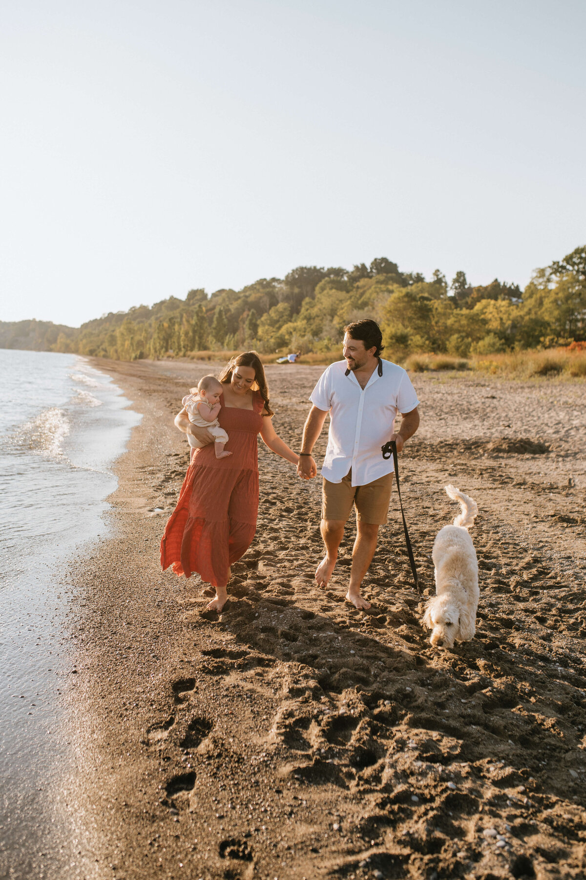Man-and-woman-walking-on-beach-holding-baby-with-dog