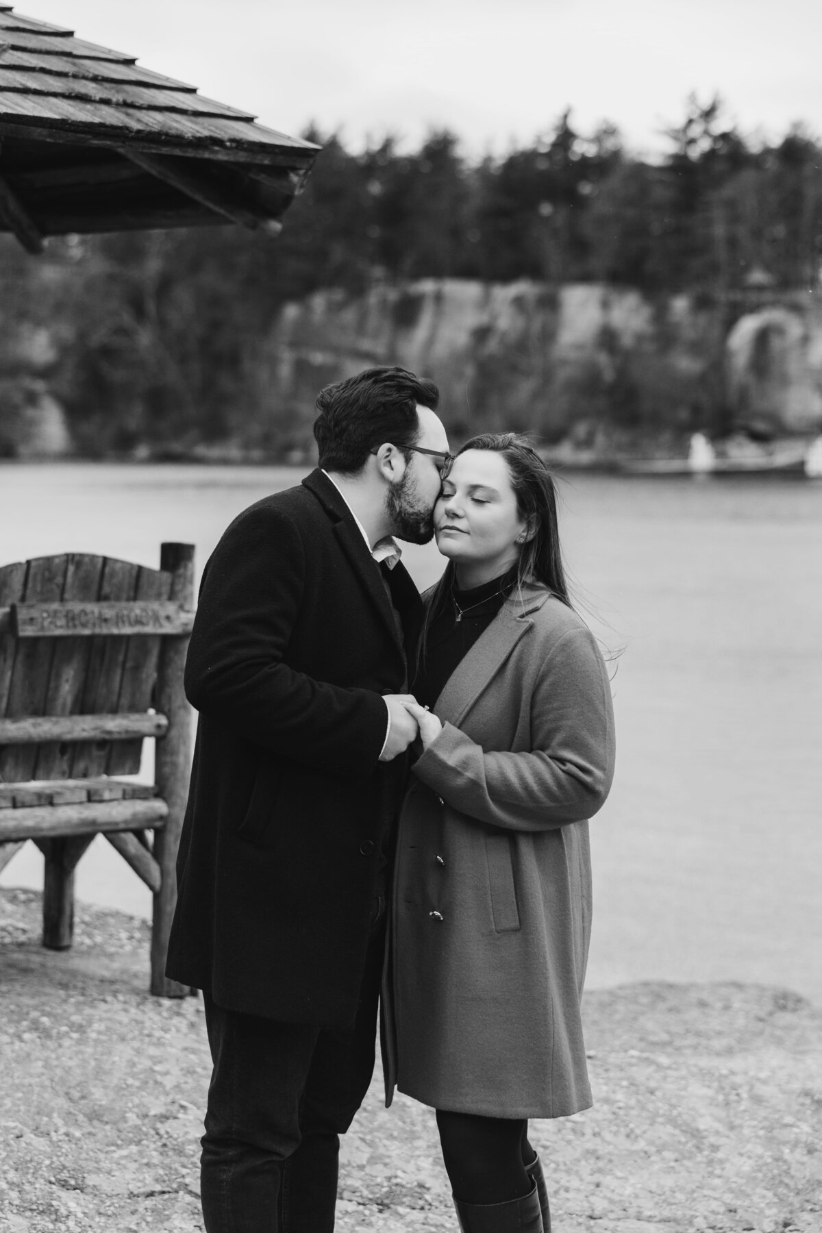 Black and white photo of a couple sharing an intimate moment by the lake at Mohonk Mountain House in Hudson Valley, holding hands and dressed in cozy winter coats.