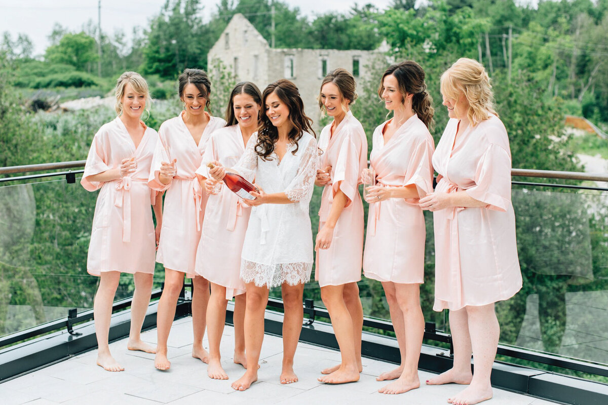 Bridal party popping champagne, wearing stylish satin robes By Catalfo, elegant wedding fashion based in Kelowna. Featured on the Brontë Bride Vendor Guide.
