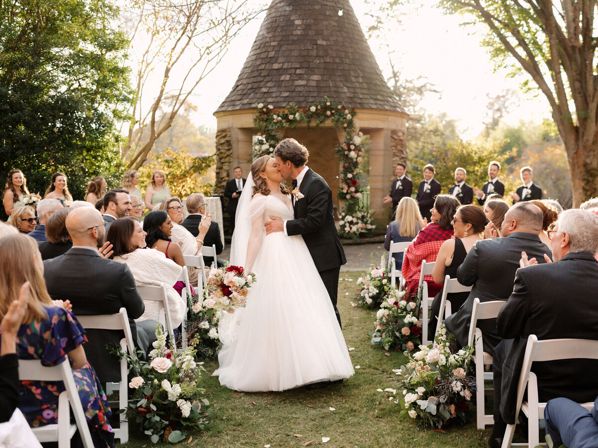 November wedding at Graylyn Estate, designed and planned by Charming Carolina — bride and groom share a kiss halfway down the floral-lined aisle after being announced as husband and wife.
