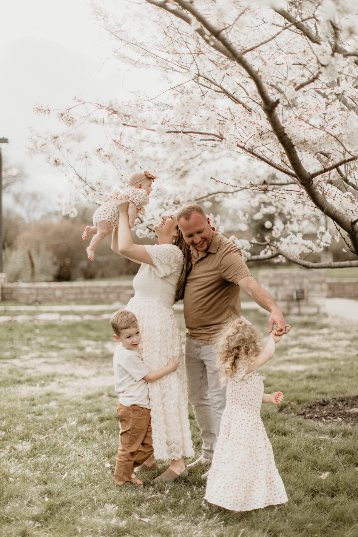 Mom and dad with 3 kids, mom holding baby up, dad spinning toddler girl. 