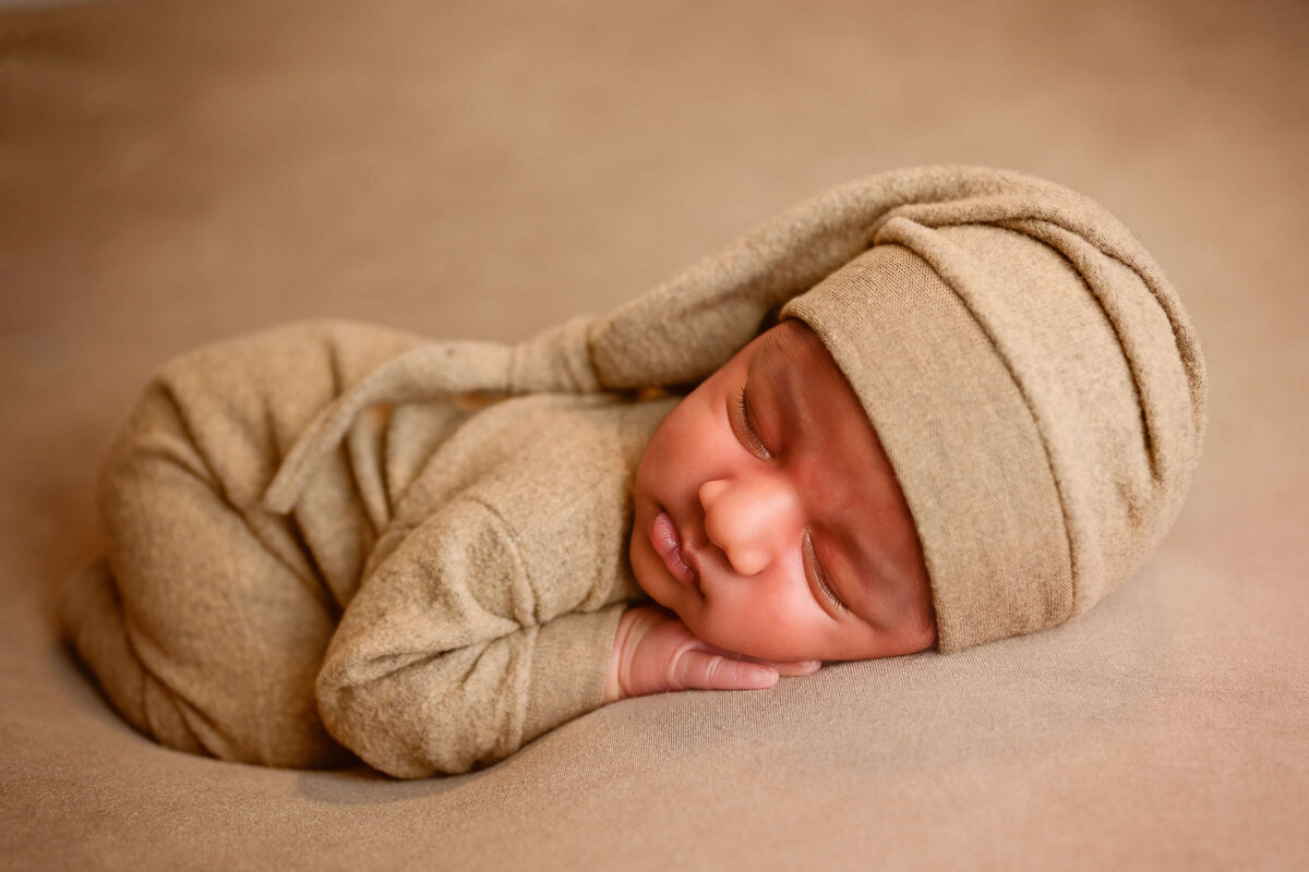 Newborn photoshoot in Kitsap County, WA. Newborn posed sleeping on his belly with hands under his cheek. Baby dressed in tan outfit with matching sleepy cap. Image by E. Benecki Photography.