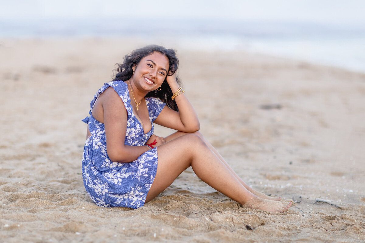 A woman sits on a sandy beach, smiling, with her right hand resting on her head. She is wearing a blue floral dress and has wavy hair. The ocean is visible in the background, with a calm and serene atmosphere.