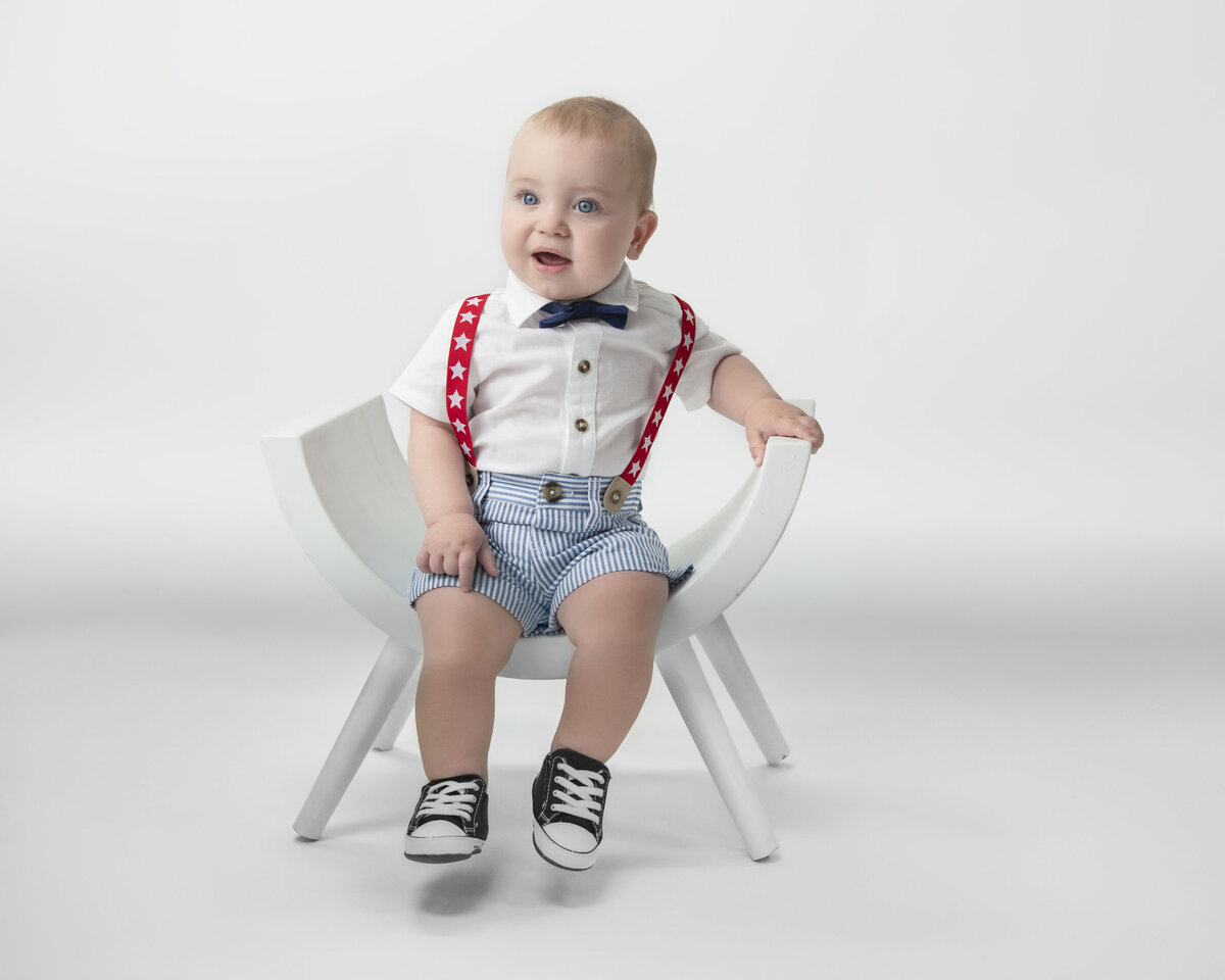 A baby in a white shirt sits in a chair in a studio with red suspenders