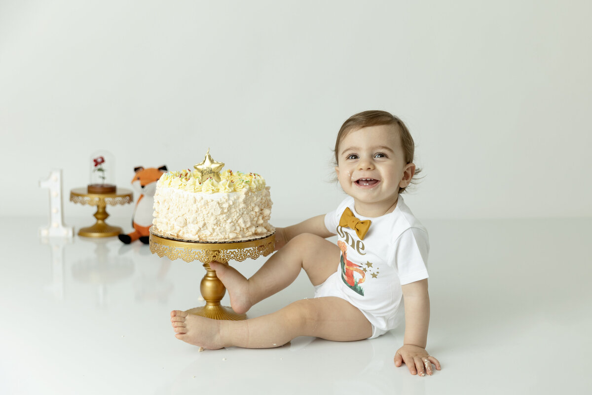 A young toddler boy smiles big before eating a yellow frosted cake in a studio wearing a gold bowtie and white onesie